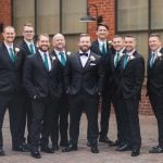 Groomsmen in black tuxedos standing in front of Savage Mill, a brick building, for a wedding portrait.