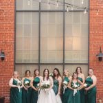 Bridesmaids in emerald dresses posing for a wedding portrait in front of the iconic brick building at Savage Mill.