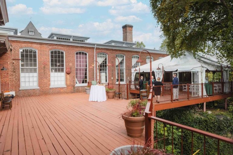 Tables and chairs are set up on a deck in front of the beautiful brick building at Savage Mill, creating the perfect setting for a wedding reception.