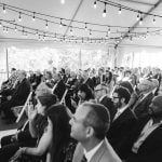 A group of people sitting in a tent watching a wedding ceremony at Savage Mill.