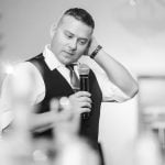 A man in a vest is holding a microphone at a wedding reception at Savage Mill.