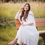 A woman in a white dress sitting on a bench in the beautiful Patuxent Research Refuge field, Maryland, indelibly capturing the essence of timeless elegance for her engagement photos.