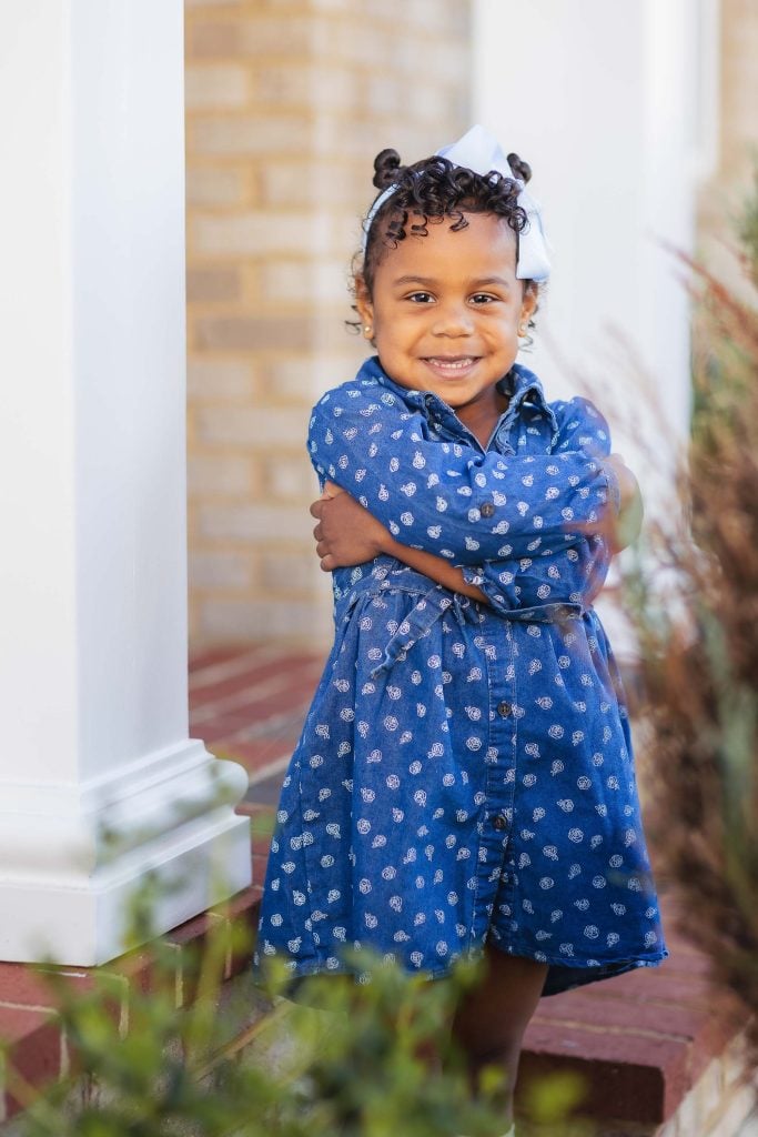 A little girl in a blue dress, standing in front of her family's home, poses for a portrait.