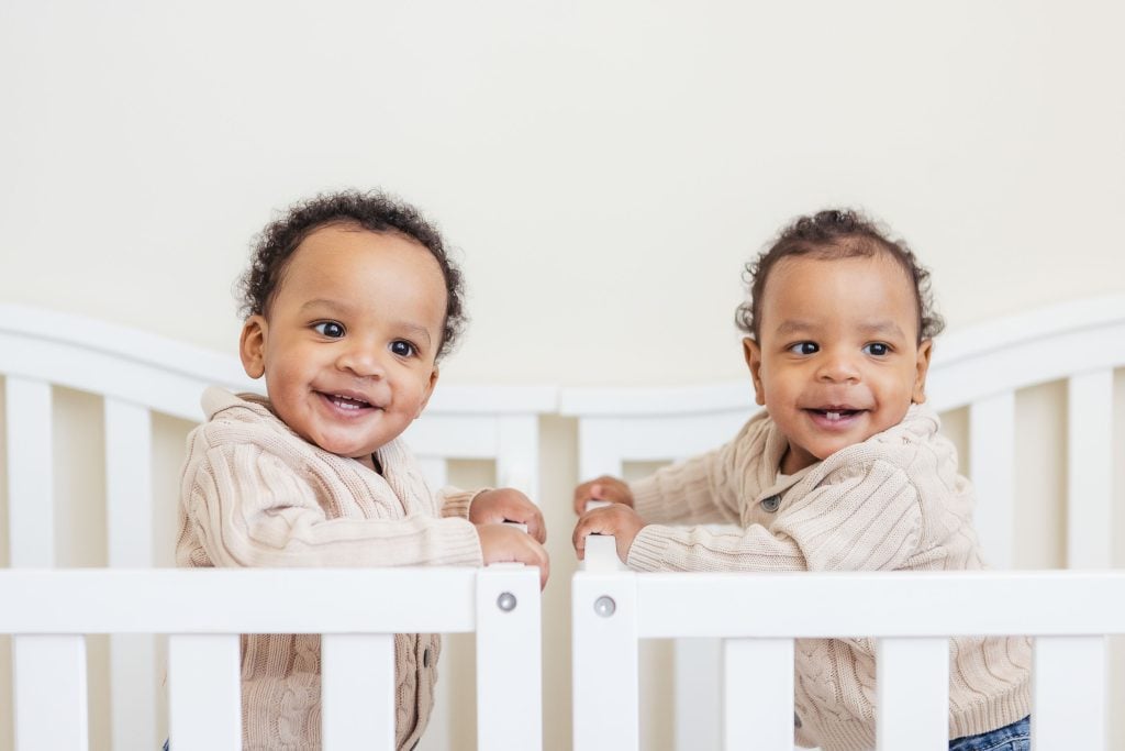 Two babies at home in a crib, gazing at each other.