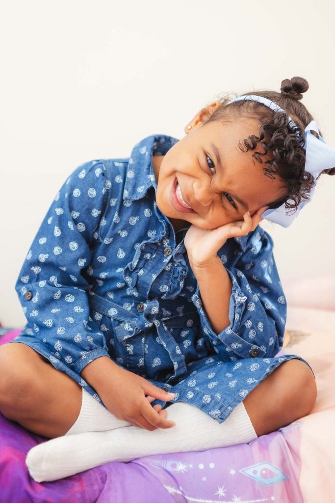 A little girl wearing a polka dot dress sitting on a bed, captured in an intimate portrait at home.