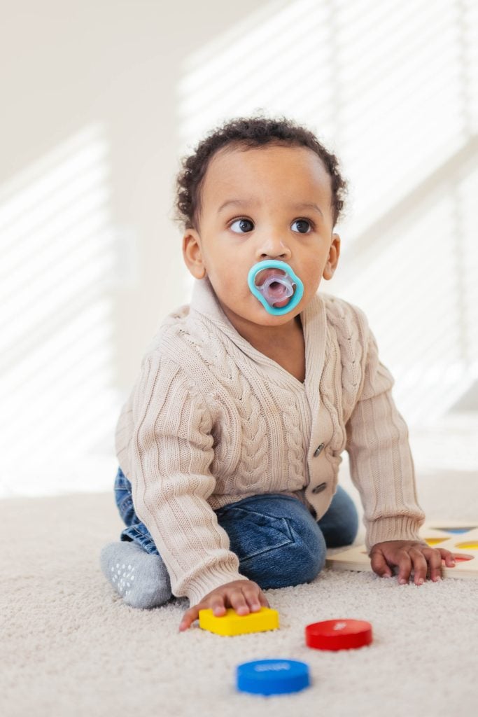 A baby with a pacifier sitting on the floor at home.