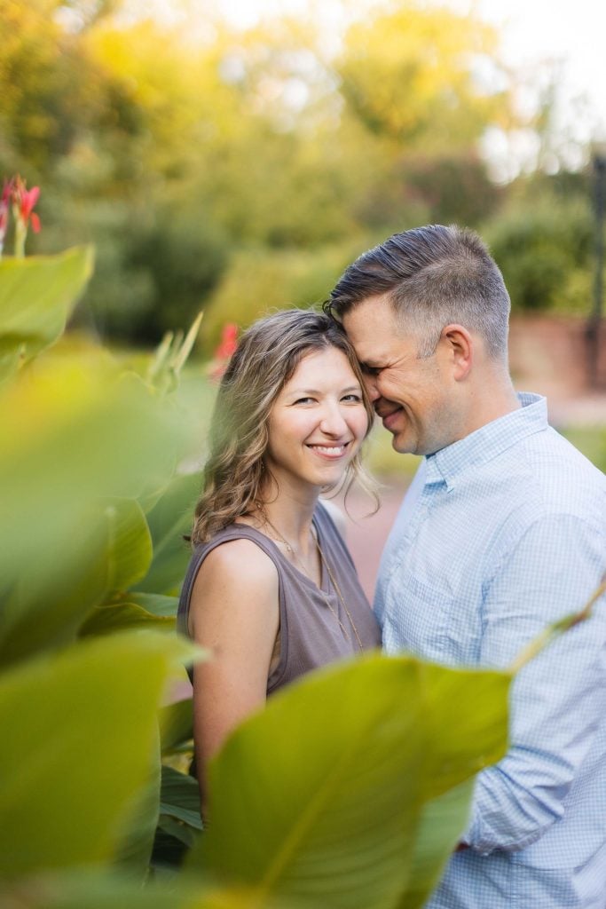 A happy engaged couple smiles in front of lush bushes during a portrait session.