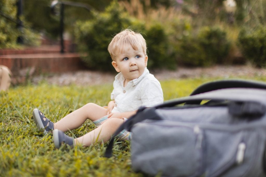 A baby sits on the grass at Quiet Waters Park, next to a bag.