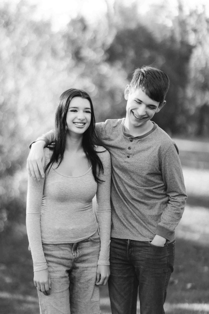 A black and white portrait of a young man with his arm around a young woman, both smiling and standing outdoors. The setting resembles the charming grounds of Belair Mansion.