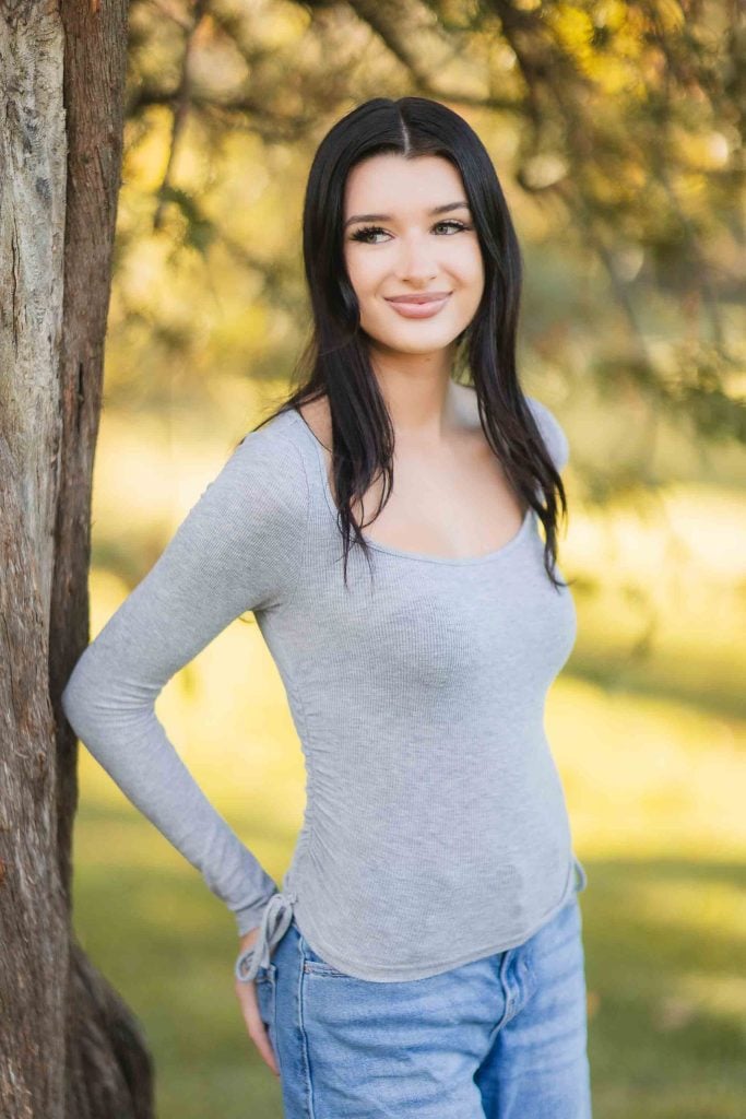 A woman with long dark hair is standing outdoors beside a tree in Bowie, Maryland, wearing a light grey longsleeve shirt and blue jeans. The background features greenery and sunlight, capturing the essence of the minisession perfectly.