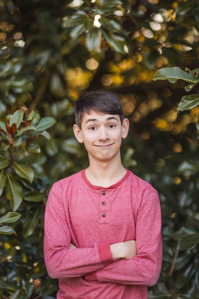 A young person with short dark hair, wearing a red longsleeve shirt, stands outdoors at Belair Mansion in Maryland with arms crossed and a playful facial expression. Green leaves create a lush background for this charming portrait.