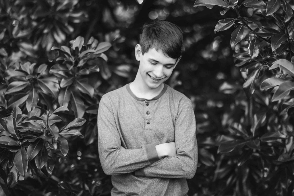 A young person stands outdoors, smiling with arms crossed, surrounded by leafy foliage. The black and white portrait captures a moment of timeless joy, reminiscent of a Bowie album cover.