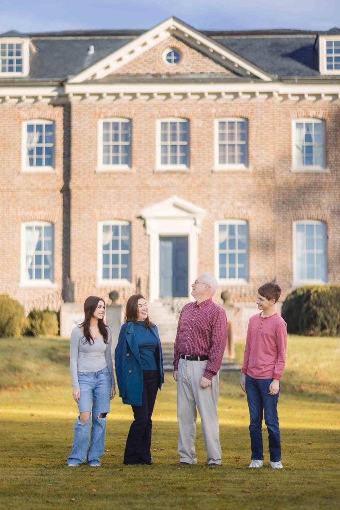 Four people stand in front of a large, twostory brick building with multiple windows and a central white door, capturing the moment as if it were a Bowieinspired portrait from a minisession.