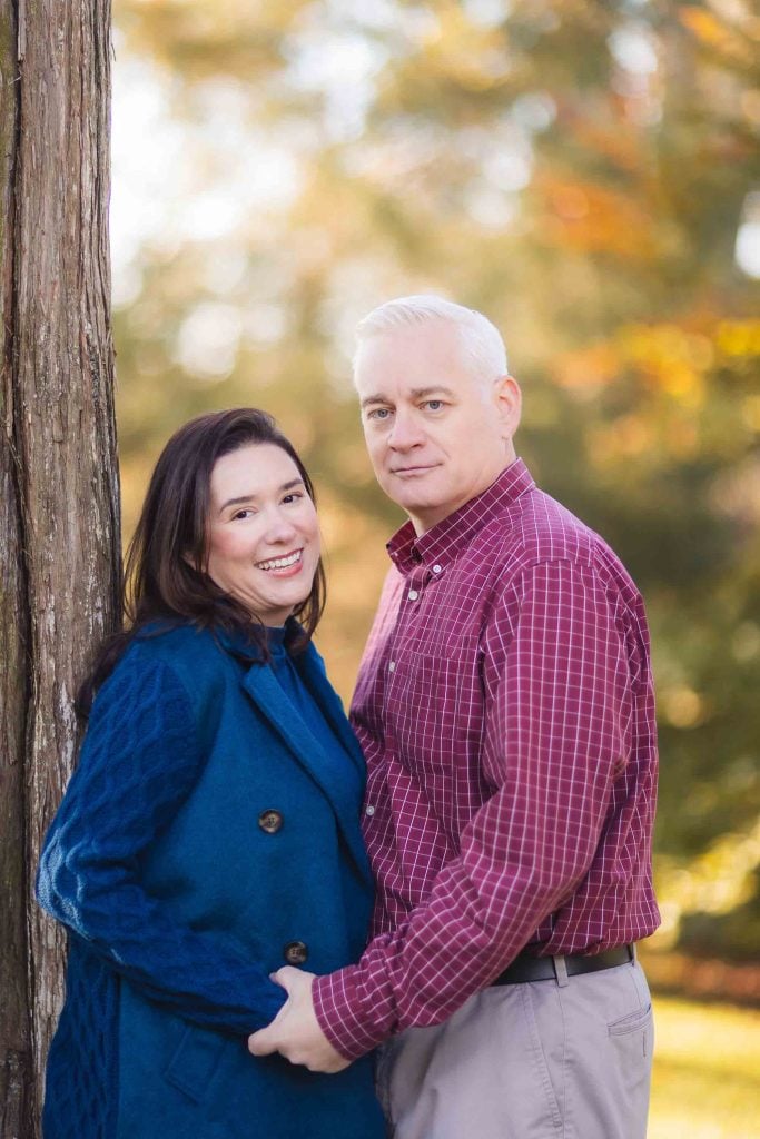 A man and woman stand outdoors holding hands, the woman smiles at the camera while the man looks ahead. They are dressed in casual autumn attire, perfect for a Bowie family minisession. Trees with fall foliage are in the background.