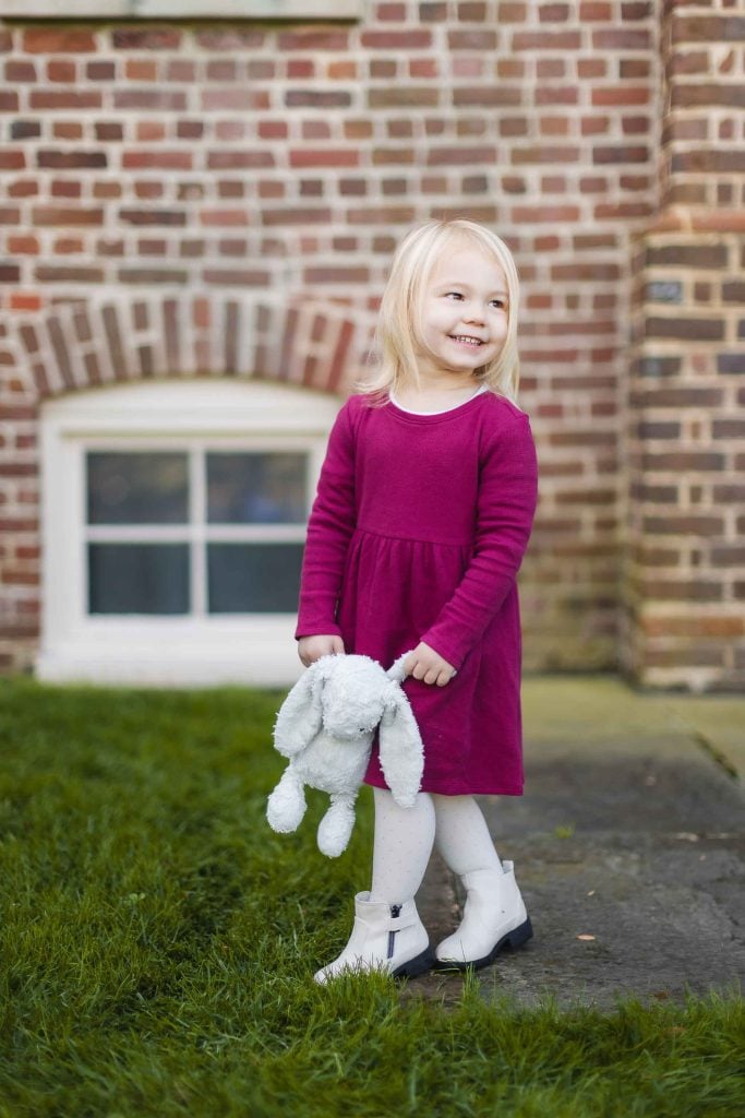 A young girl in a maroon dress stands outside in front of a brick building, holding a plush white rabbit. She is smiling and looking to her left, capturing a perfect moment for this family portrait minisession.