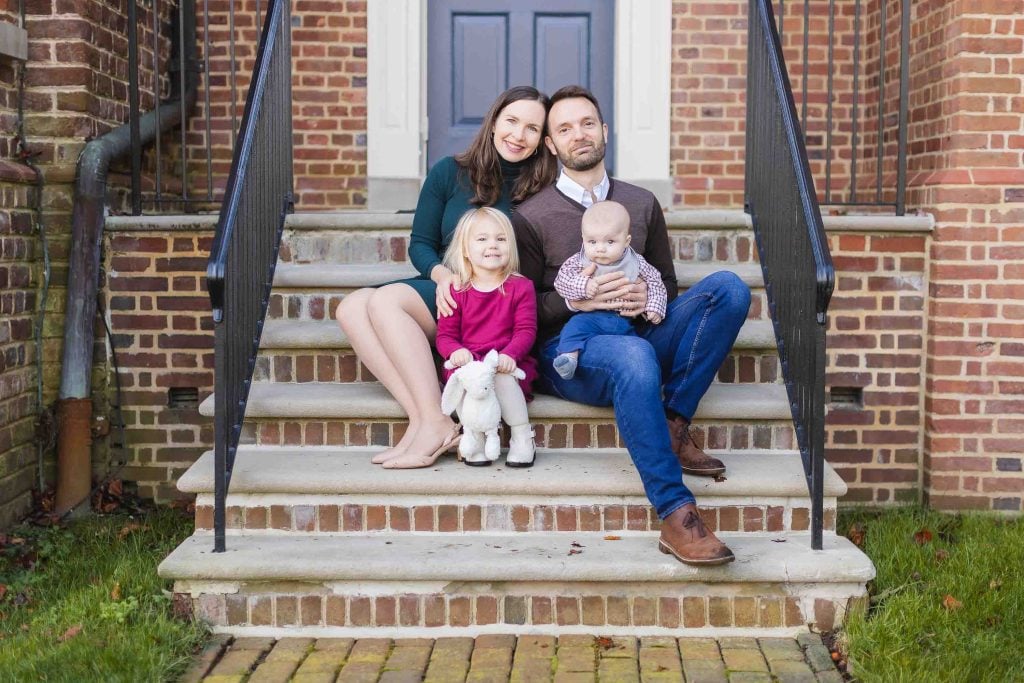 A family of four sits on the front steps of a brick house in Maryland. The woman holds a young girl with a toy, while the man cradles a baby. The scene captures the warmth and togetherness reminiscent of moments at places like Belair Mansion.