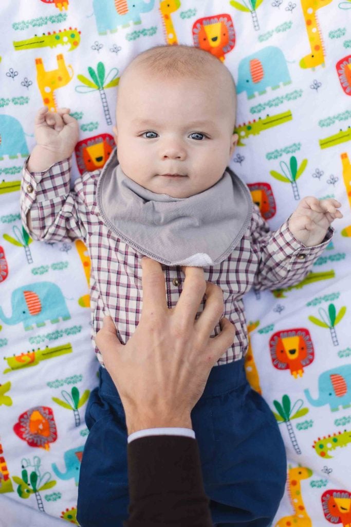 During a charming minisession in Maryland, a baby wearing a checkered shirt and a bib lies on a colorful blanket with animal prints as an adult hand gently touches the baby's chest, capturing the perfect portrait moment.