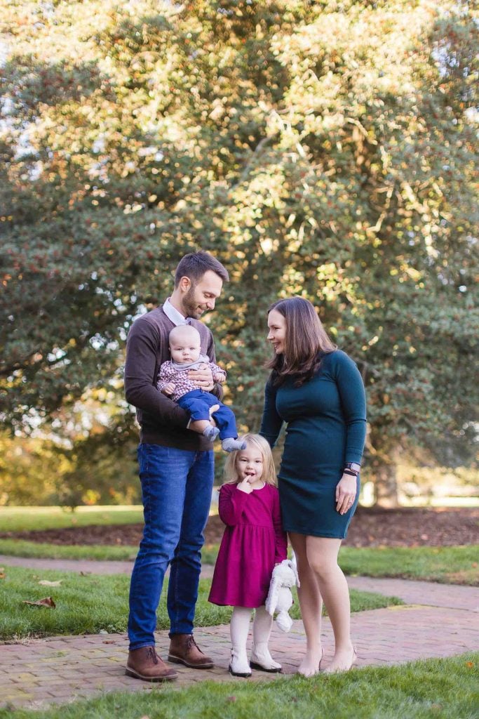 A man and woman stand on a walkway in a park in Bowie, Maryland, with the man holding a baby and the woman next to a young girl in a red dress. Trees and greenery provide a beautiful backdrop for this delightful minisession.