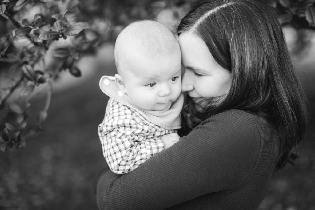 A woman holds a baby close, with the baby's face resting against her cheek, in a tender embrace. The black and white portrait captures this intimate moment against a blurred background of leafy branches. This heartwarming minisession took place in Maryland.