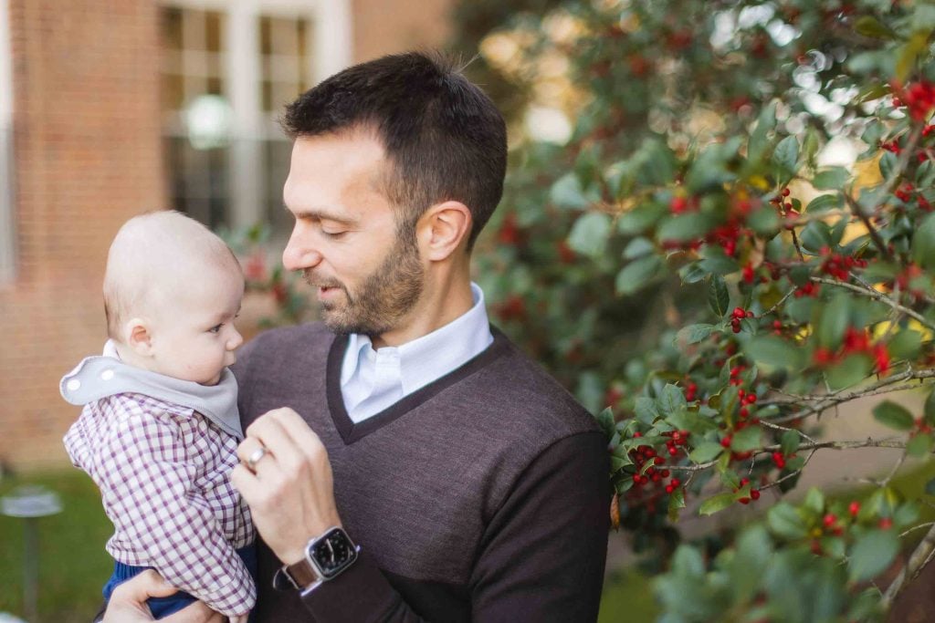 In this charming portrait taken in Bowie, Maryland, a man holds a baby near a shrub with red berries. The man is smiling at the baby and wearing a brown sweater, while the baby has on a checkered shirt and bib. A brick building is visible in the background.