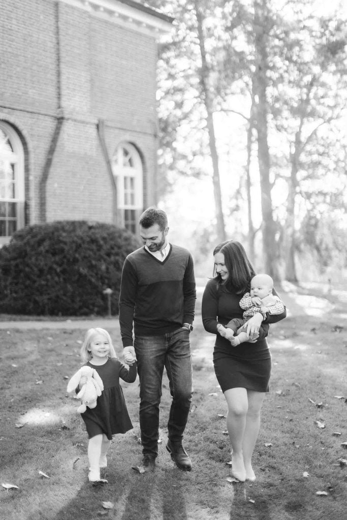 A family of four, with a mother holding a baby and a father holding a young child's hand, strolls on the grassy area beside the historic Belair Mansion in Bowie, surrounded by trees and a charming brick building.