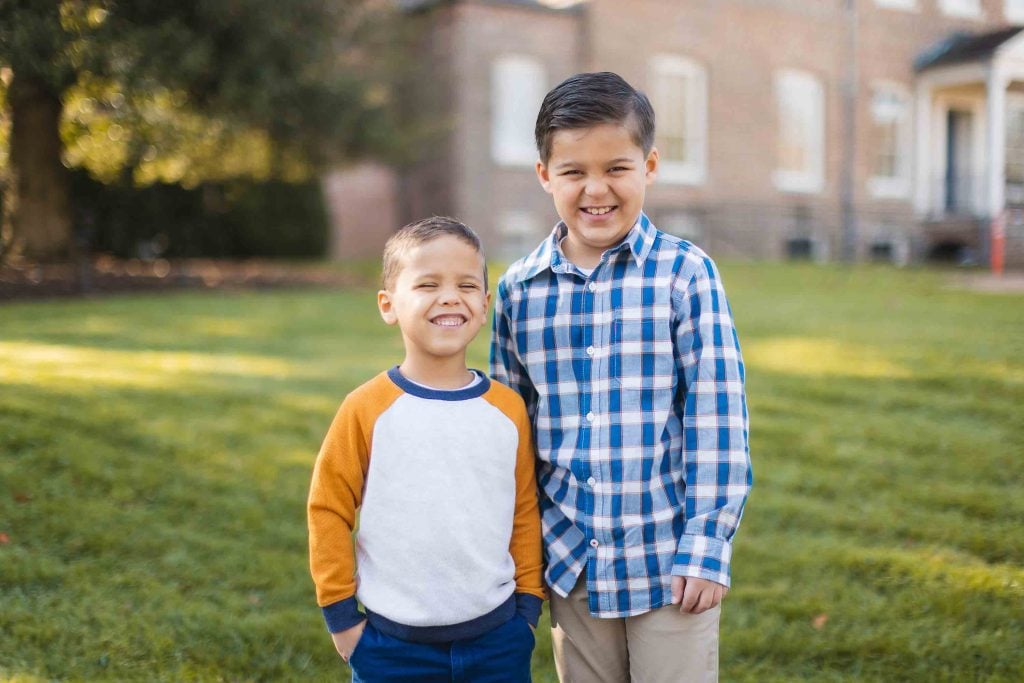 Two young boys stand close together and smile in this charming portrait taken outdoors on a grassy area, with a building and trees in the background during their minisession.