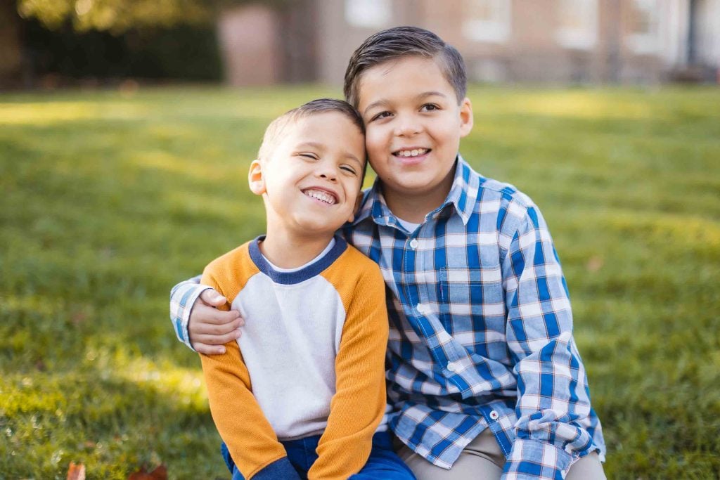 Two young boys sitting on the grass at Belair Mansion in Bowie, the older one wearing a blue plaid shirt and the younger one in a yellow and white shirt, with the older boy's arm around the younger during a minisession.