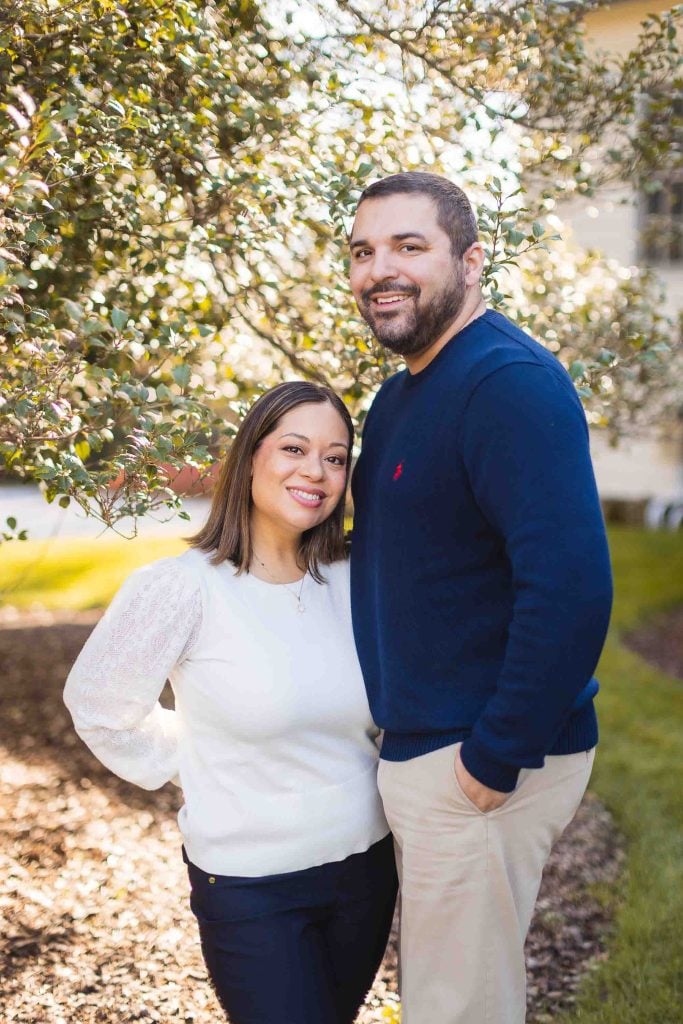 A man and woman stand closely together outdoors near a bush at Belair Mansion, smiling at the camera. The man wears a navy sweater and beige pants, while the woman wears a white sweater and dark pants. This lovely moment was captured during their minisession.