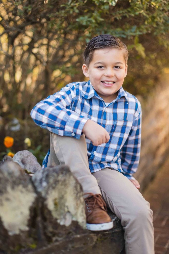 A boy with neatly combed hair and wearing a blue checkered shirt and khaki pants sits on a stone wall, smiling at the camera in what feels like a delightful minisession portrait.