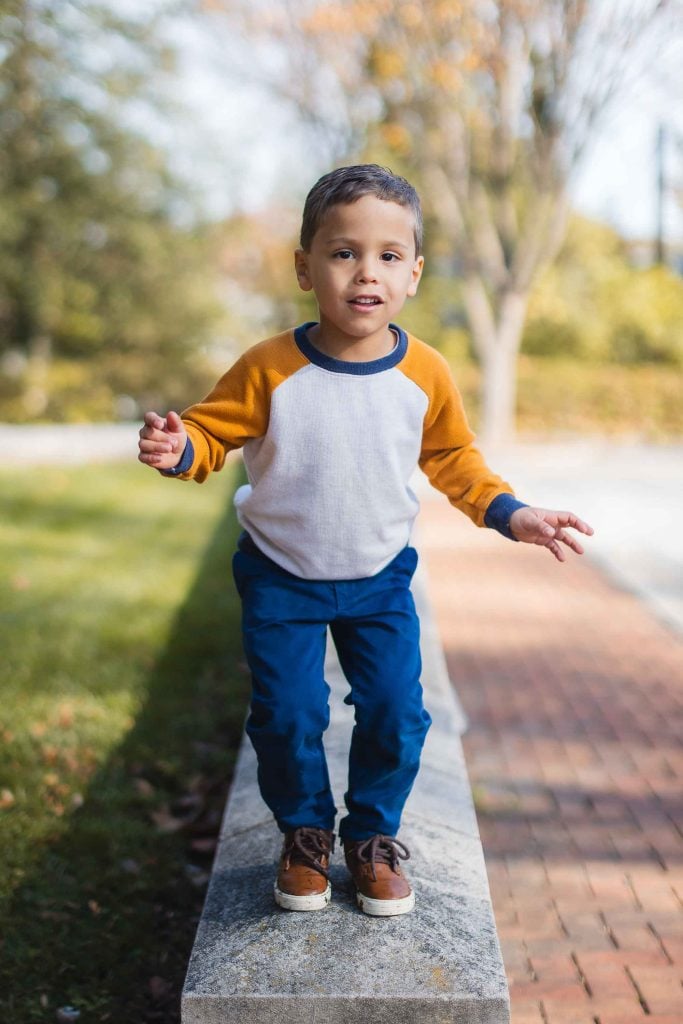 A young boy in a white and orange longsleeve shirt and blue pants stands on a low concrete ledge outdoors, appearing to balance himself with extended arms, reminiscent of carefree days spent exploring the grounds of Belair Mansion.