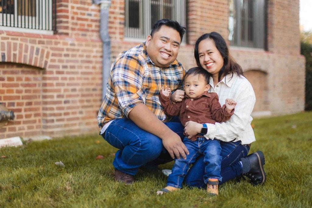 A smiling couple kneels in the grass outside a brick building, holding a toddler between them. This charming family portrait, dressed in casual plaid and denim outfits, captures a candid moment of joy.