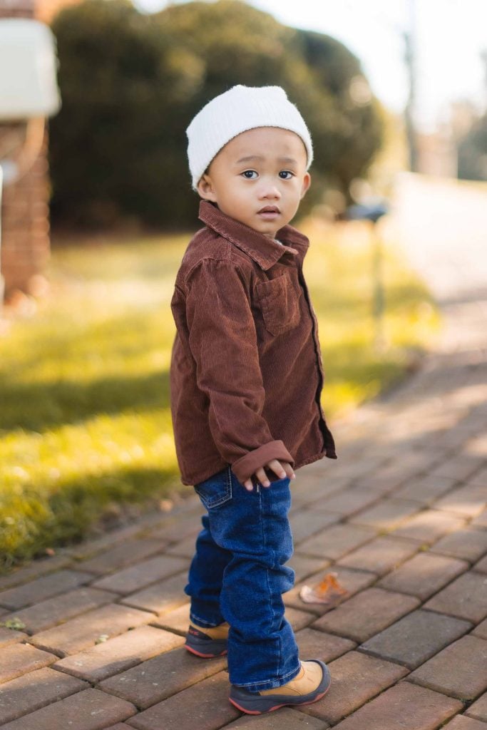 A young child wearing a white beanie, brown jacket, blue jeans, and brown shoes stands on a brick path at Belair Mansion, looking over their shoulder. The minisession captures the essence of curiosity with a grassy area and blurred background visible.