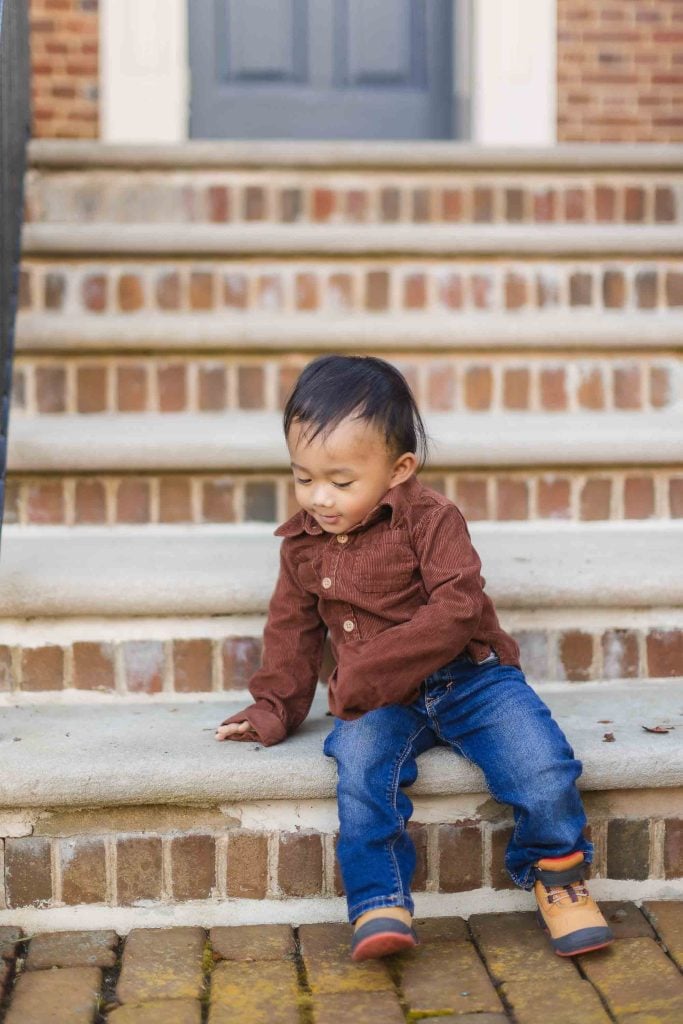 A young child in a brown shirt and blue jeans sits on a set of outdoor steps at the historic Belair Mansion, enjoying a sunny day with family in Bowie.