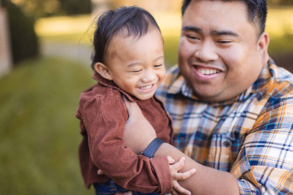 A joyful adult and a child smile, with the child held in the adult's arms. The adult wears a plaid shirt and the child wears a brown outfit. They are outdoors at Belair Mansion, Maryland, with blurred greenery in the background during their minisession.