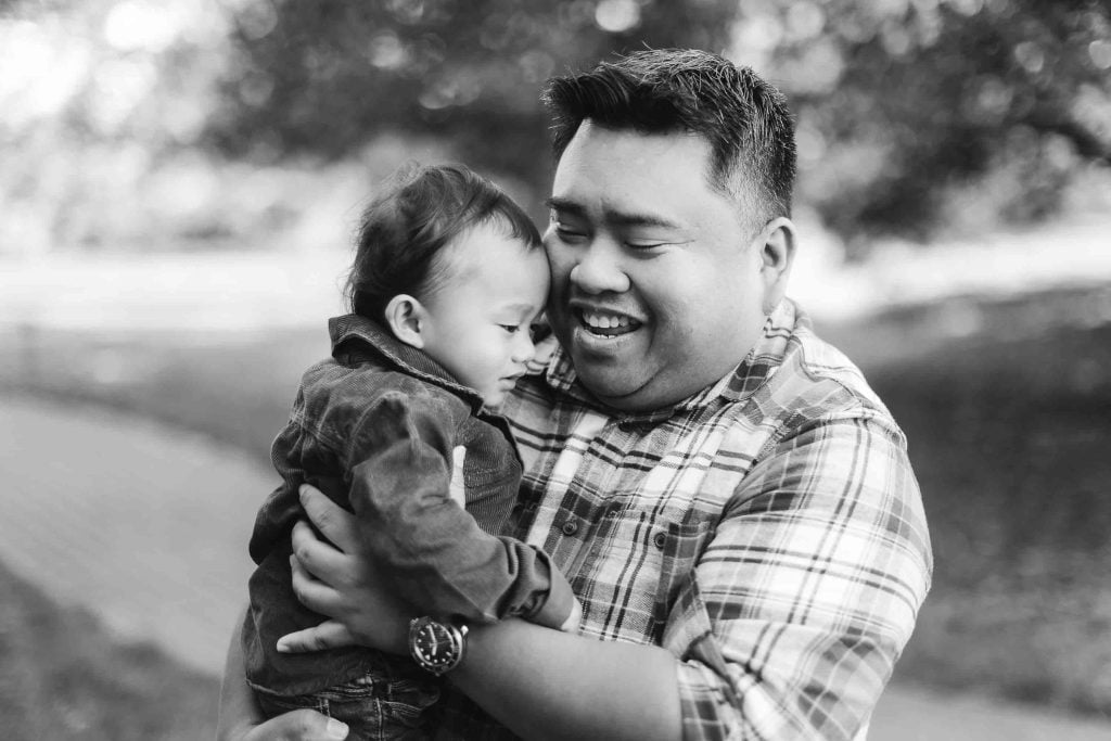 A smiling man in a plaid shirt holds a baby in his arms outdoors during a portrait minisession. Both appear to be content and happy. The background shows blurred trees and a pathway, capturing the serene beauty of Maryland.