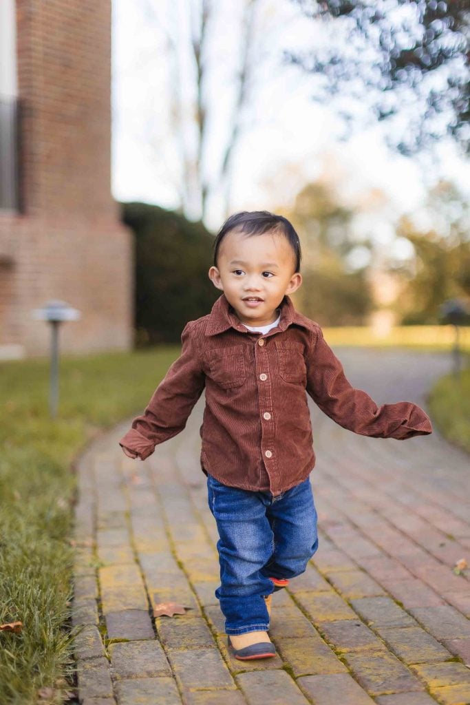 A young child with short dark hair walks down a brick path in Bowie, Maryland. The child is wearing a brown shirt and blue jeans. Trees and a building are visible in the background, showcasing the charm of this familyfriendly community.