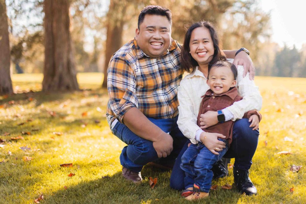 A smiling family of three sits and kneels together on grass in a park, with trees and sunlight in the background. This charming portrait captures a man in a plaid shirt, a woman in a cream shirt, and a child in brown and blue jackets during their minisession.