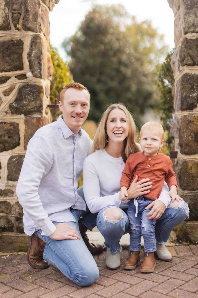 A family of three poses for a minisession photo outdoors in front of a stone archway in Maryland. The man, woman, and young child all smile warmly, dressed in casual clothing.