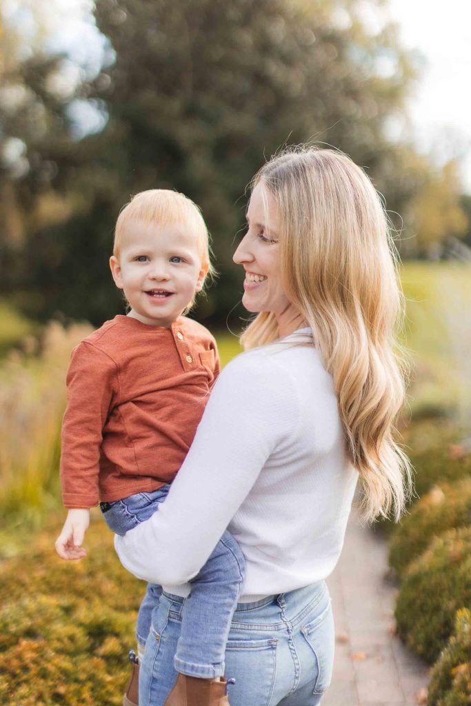 A woman with blonde hair stands outdoors, holding a young child wearing an orange shirt. The background shows greenery and a pathway, creating the perfect setting for a minisession portrait.