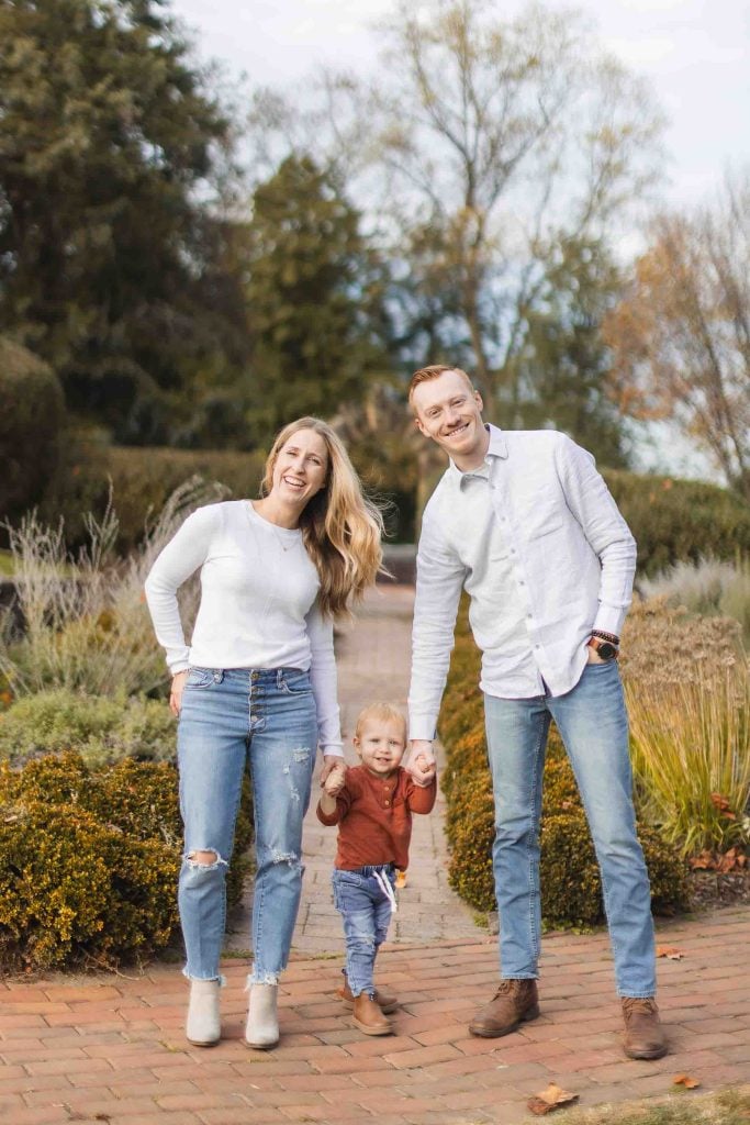 A smiling family of three, parents and a young child, holding hands and standing on a brick pathway in Belair Mansion's park in Bowie, Maryland with trees and bushes in the background.