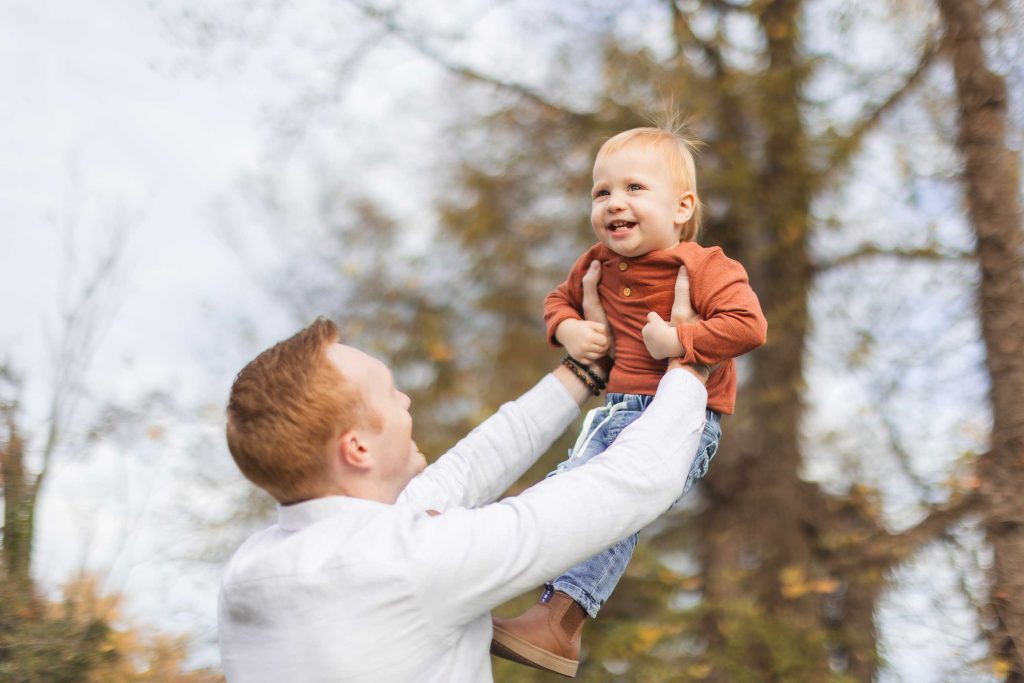 During a delightful family minisession at Belair Mansion, an adult in a white shirt lifts a smiling toddler in an orange shirt and blue jeans against the backdrop of lush trees.