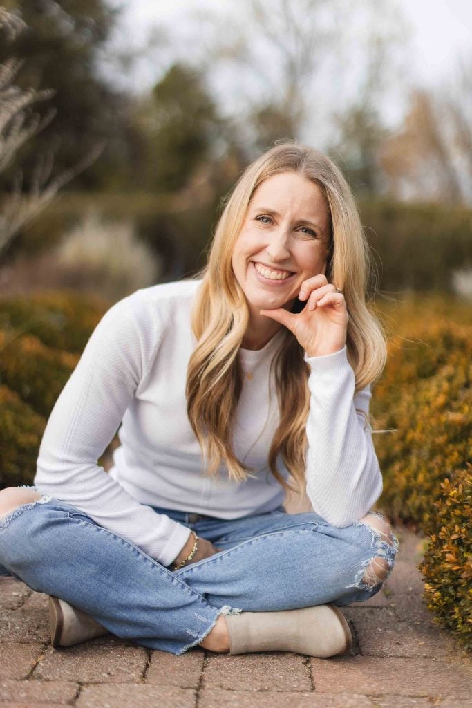 A woman with long blonde hair wearing a white sweater and ripped jeans sits crosslegged on a brick path outdoors, smiling and resting her chin on her hand. Surrounded by shrubs and trees, she exudes warmth as if waiting for family at the picturesque Belair Mansion.