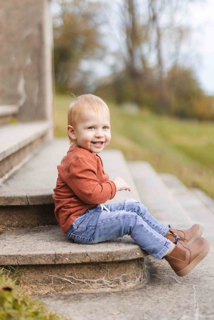 A young child wearing a red longsleeve shirt, blue jeans, and brown ankle boots sits smiling on the outdoor stone steps of Belair Mansion with a blurred green field and trees in the background.