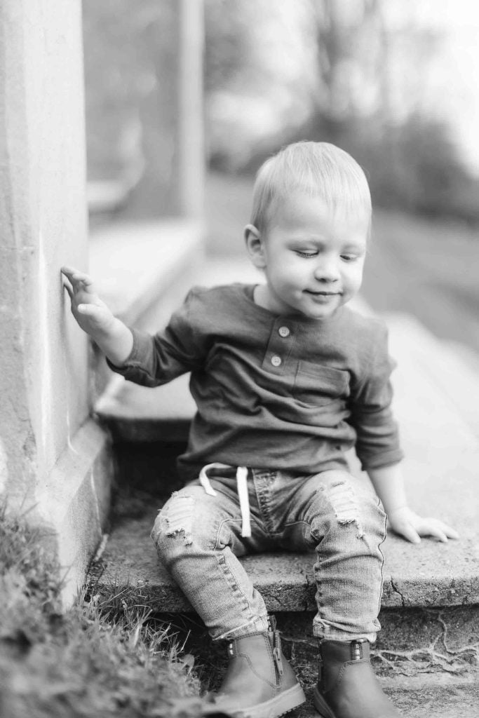 A black and white portrait captures a young child in Maryland sitting on outdoor steps. The child, donning a longsleeve shirt, ripped jeans, and boots, looks down with one hand resting on the step—a perfect moment from a minisession.