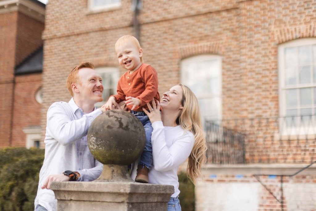 A man and a woman hold up a smiling toddler standing on a stone pedestal in front of a brick building during a minisession in Bowie, Maryland.