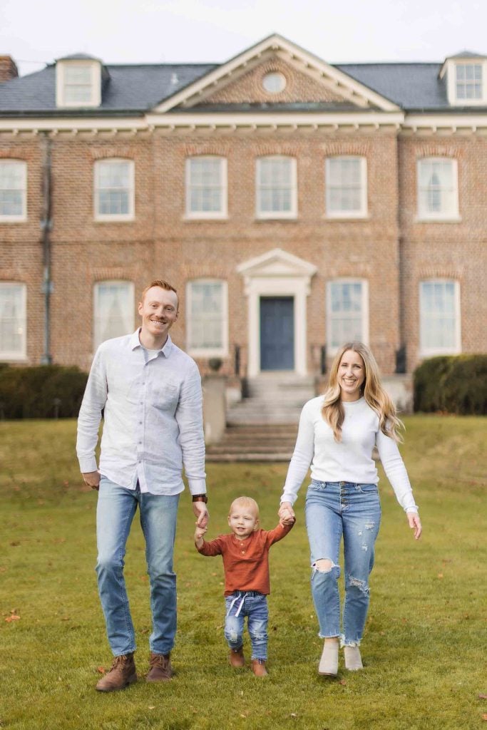 A family walks hand in hand with a young child between them on a lawn in front of a large brick building, capturing the perfect moment during their Maryland minisession.