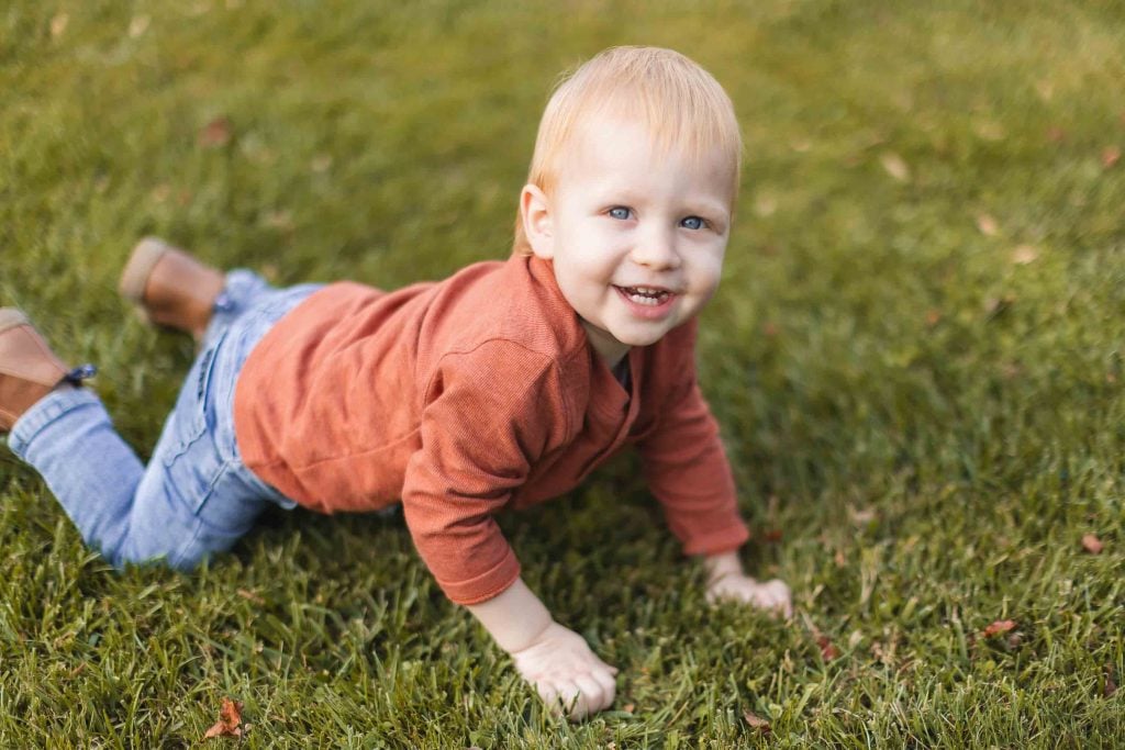 A young child wearing a red longsleeve shirt and blue jeans is crawling on the grass, looking at the camera and smiling, at a family picnic near the historic Belair Mansion in Maryland.