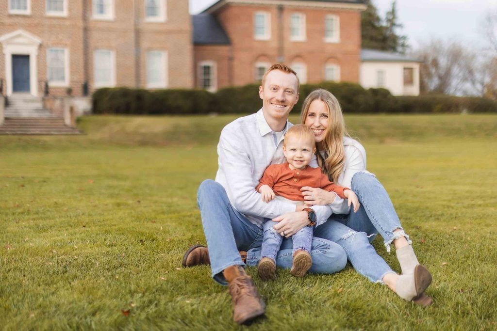 A man, woman, and small child sit on grass in front of Belair Mansion in Bowie, Maryland, all smiling at the camera.