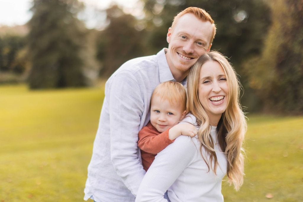 A smiling man, woman, and young child stand together in an outdoor setting at what appears to be a minisession. The child is between the adults, being held by the woman. Trees and grass are visible in the background, capturing a serene moment perhaps near Belair Mansion in Maryland.