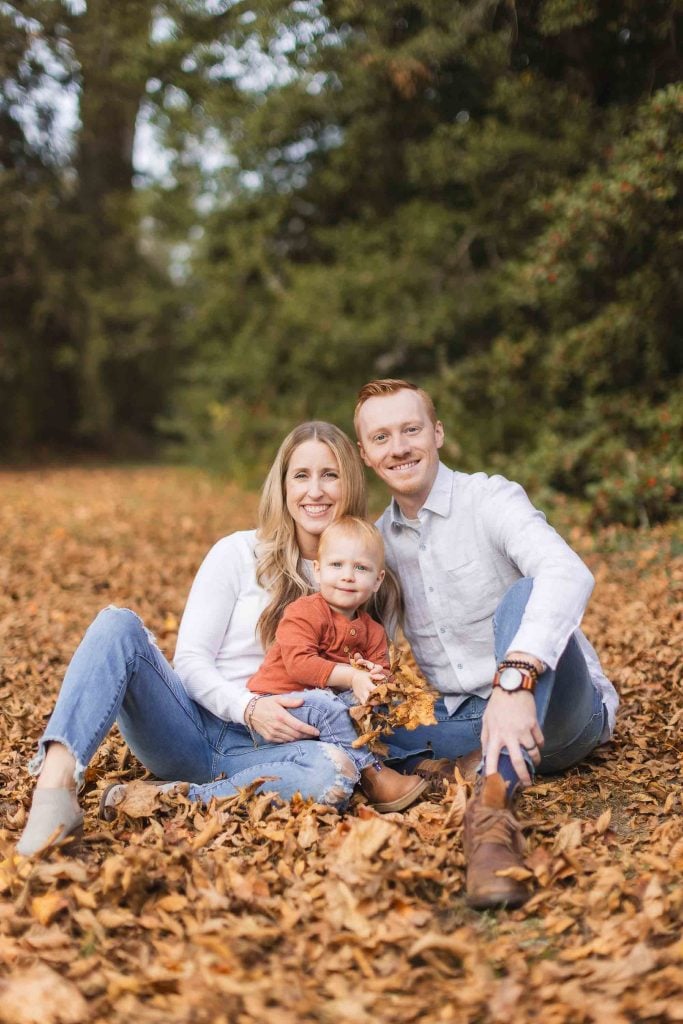 A family of three sits on the ground covered with autumn leaves, smiling at the camera. The parents are dressed in casual white tops, and the child is wearing a rustcolored shirt. Trees are in the background, creating a perfect portrait reminiscent of a scene near Belair Mansion in Bowie.